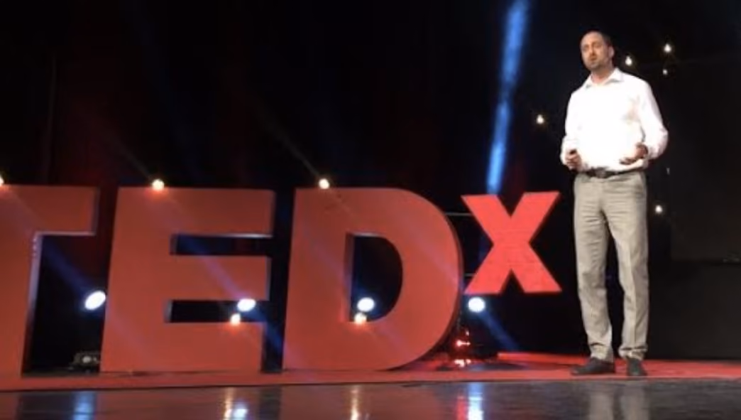 Man in white shirt and gray pants giving a TEDx talk on stage with large red TEDx letters.