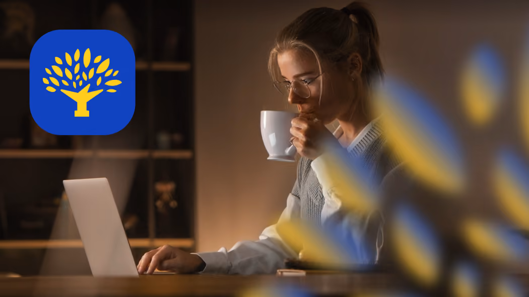 Woman drinking coffee while working on a laptop in a dimly lit room with a tree logo in the corner.
