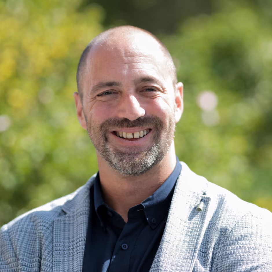 Smiling bald man with a beard wearing a dark shirt and light gray jacket outdoors.