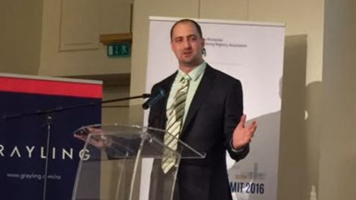 Man in suit speaking at a clear podium with a microphone and conference banners behind him.