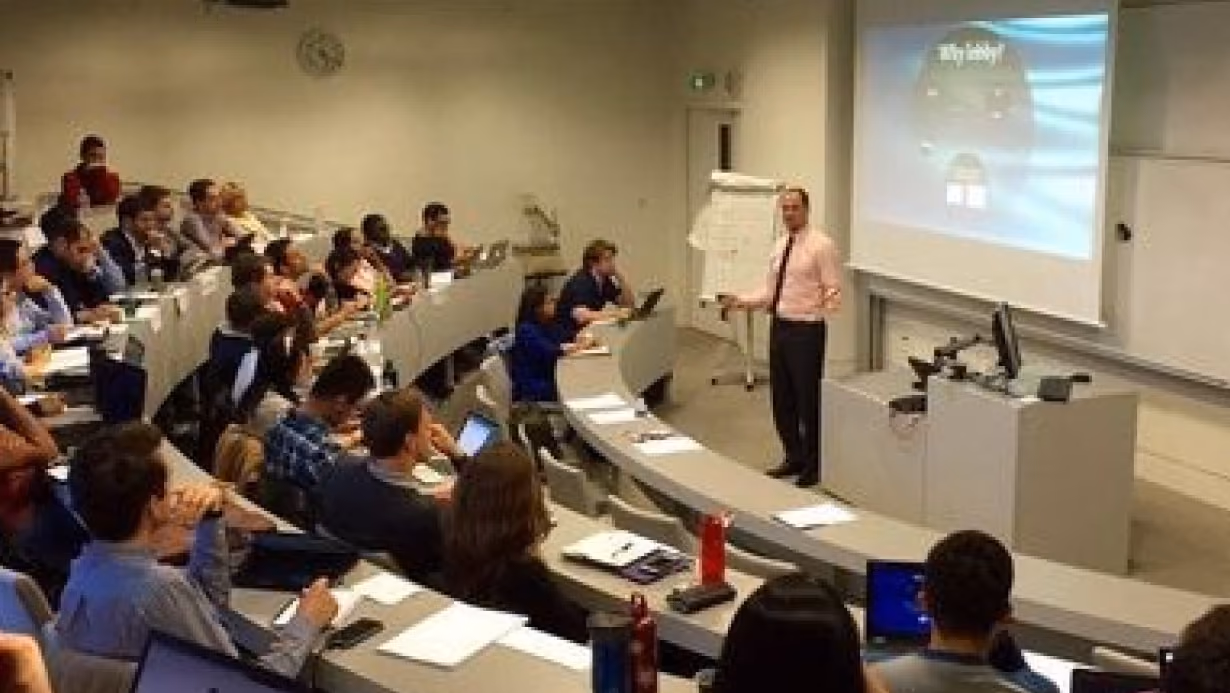 Professor giving lecture to a diverse group of students seated in tiered classroom with laptops and notes.