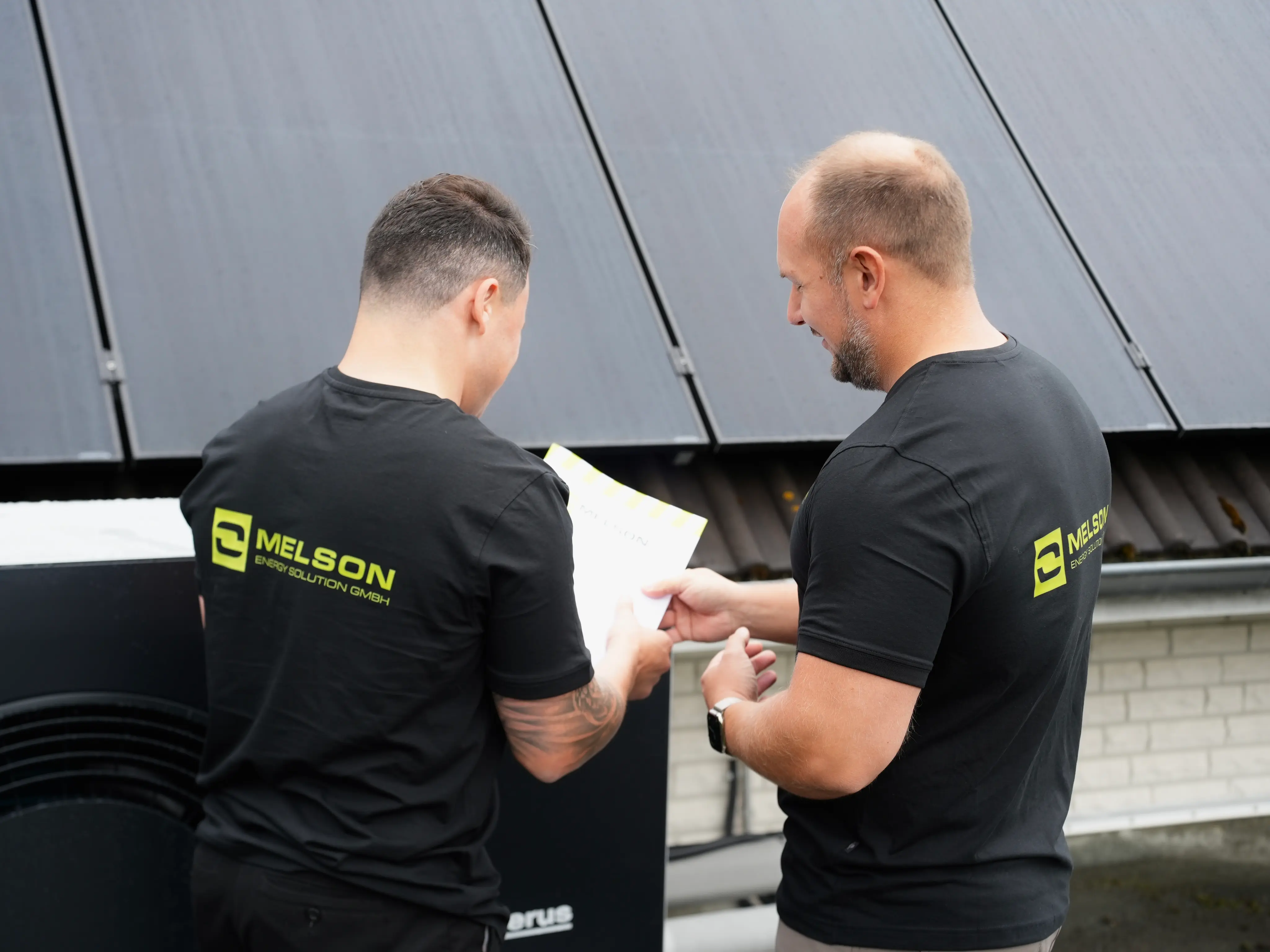 Zwei Melson Home Techniker im Gespräch über Unterlagen vor einer installierten Wärmepumpe und Photovoltaikanlage auf dem Dach eines Wohnhauses.