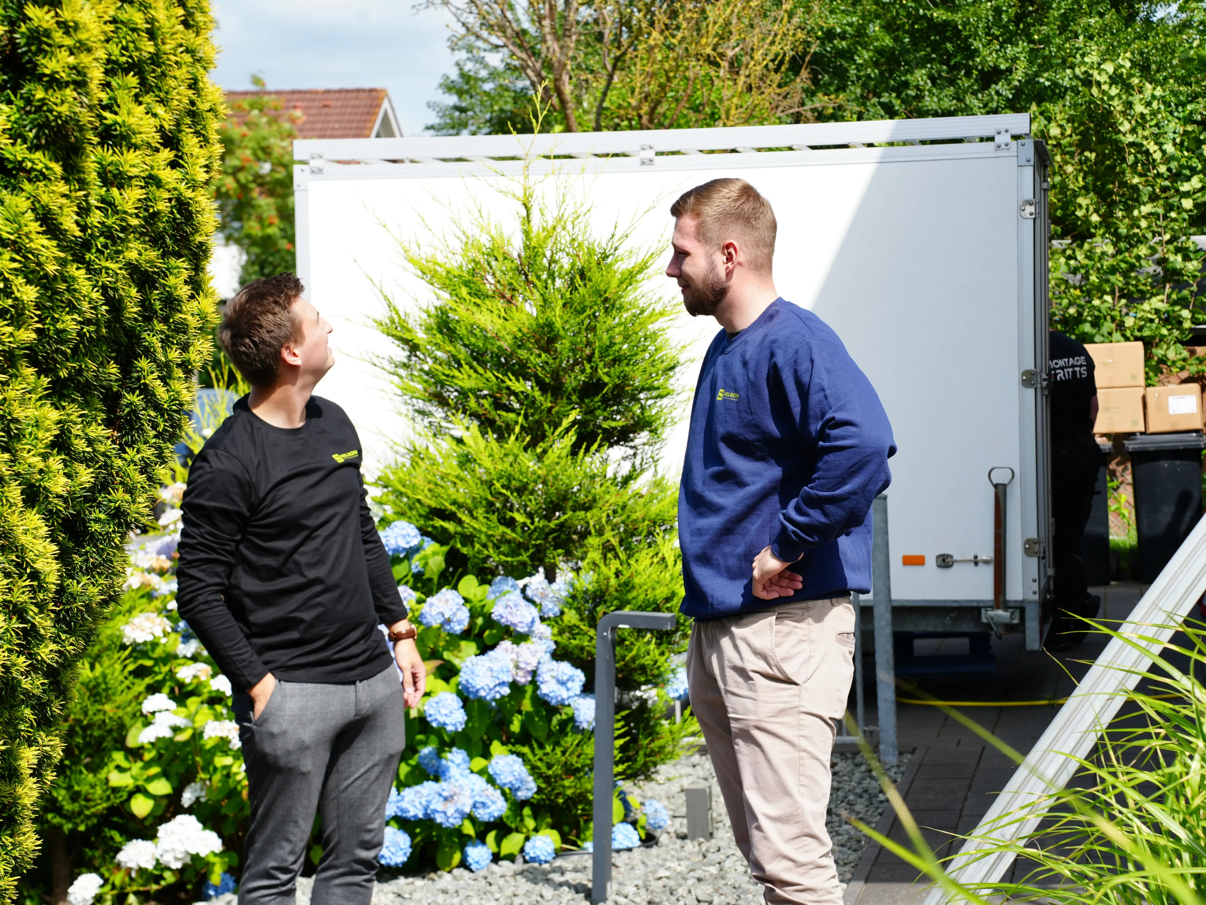 Zwei Melson Home Mitarbeiter im Gespräch vor einem Kundenhaus, umgeben von blühenden Pflanzen – im Hintergrund ein Montagefahrzeug für die Installation von Photovoltaik oder Wärmepumpe.