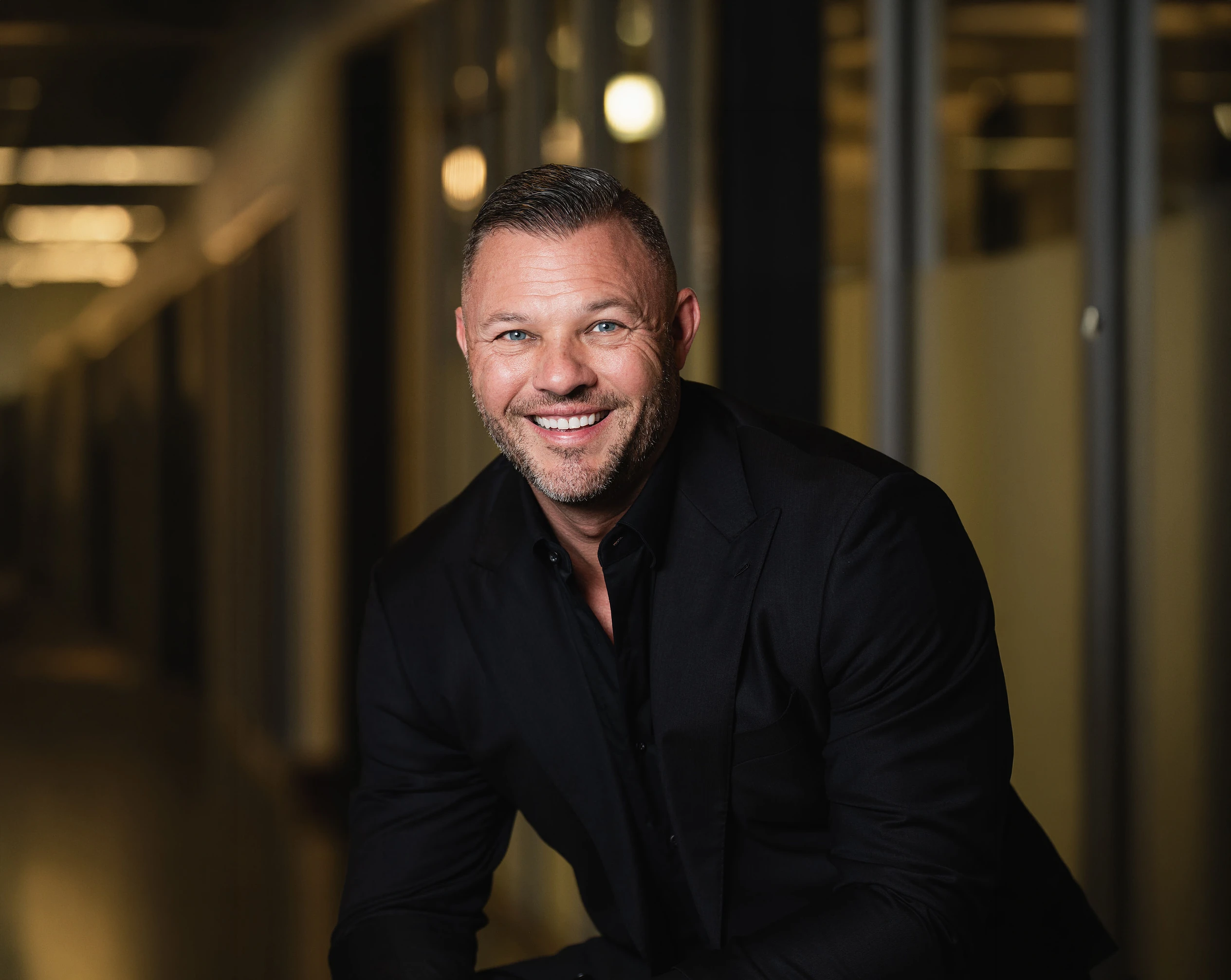 Smiling man with short hair and beard wearing a black suit in a dimly lit modern hallway.