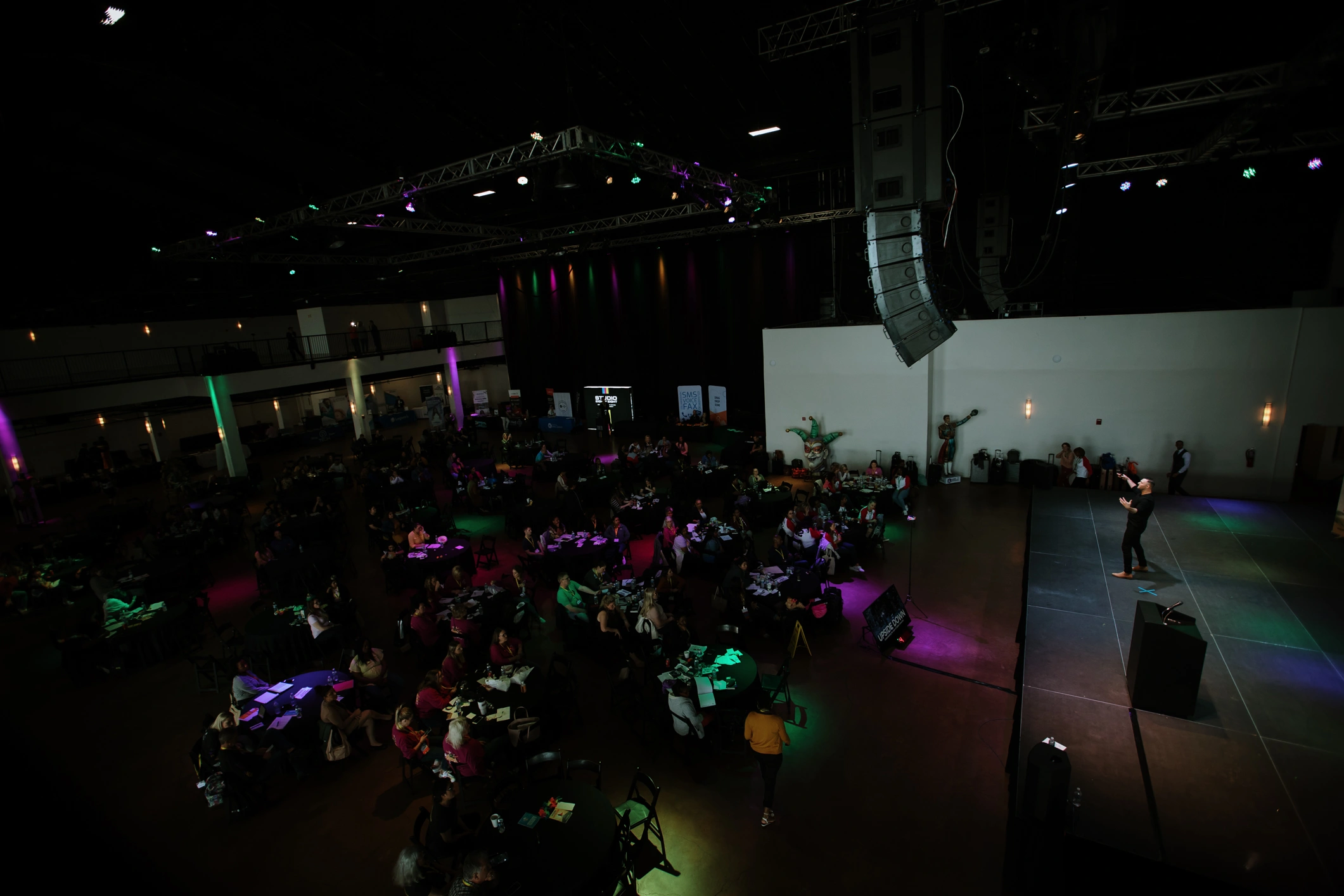 Speaker on stage addressing an audience seated at round tables in a dimly lit large event hall.