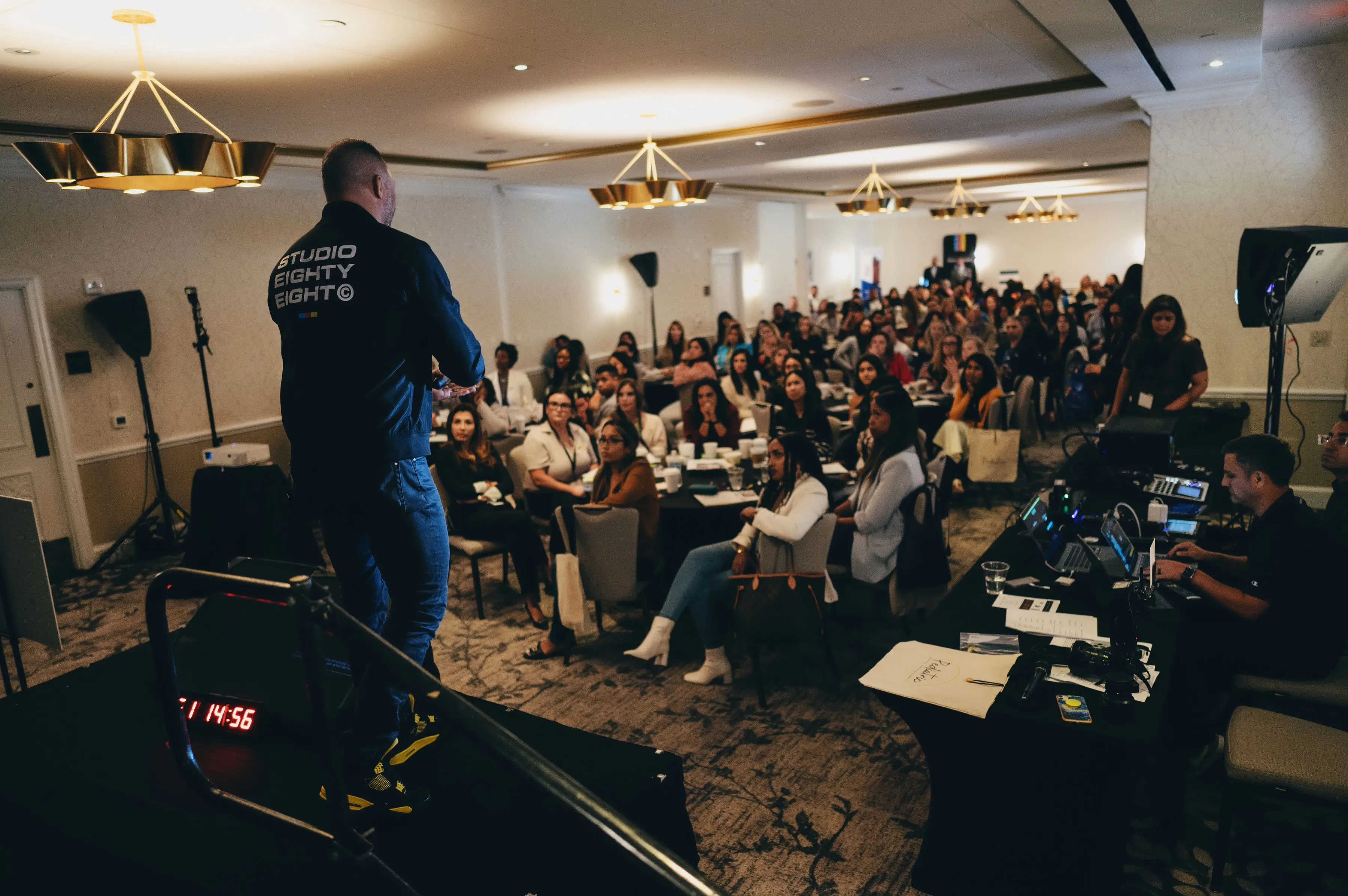 Speaker in Studio Eighty Eight jacket addresses a large seated audience in a conference room.
