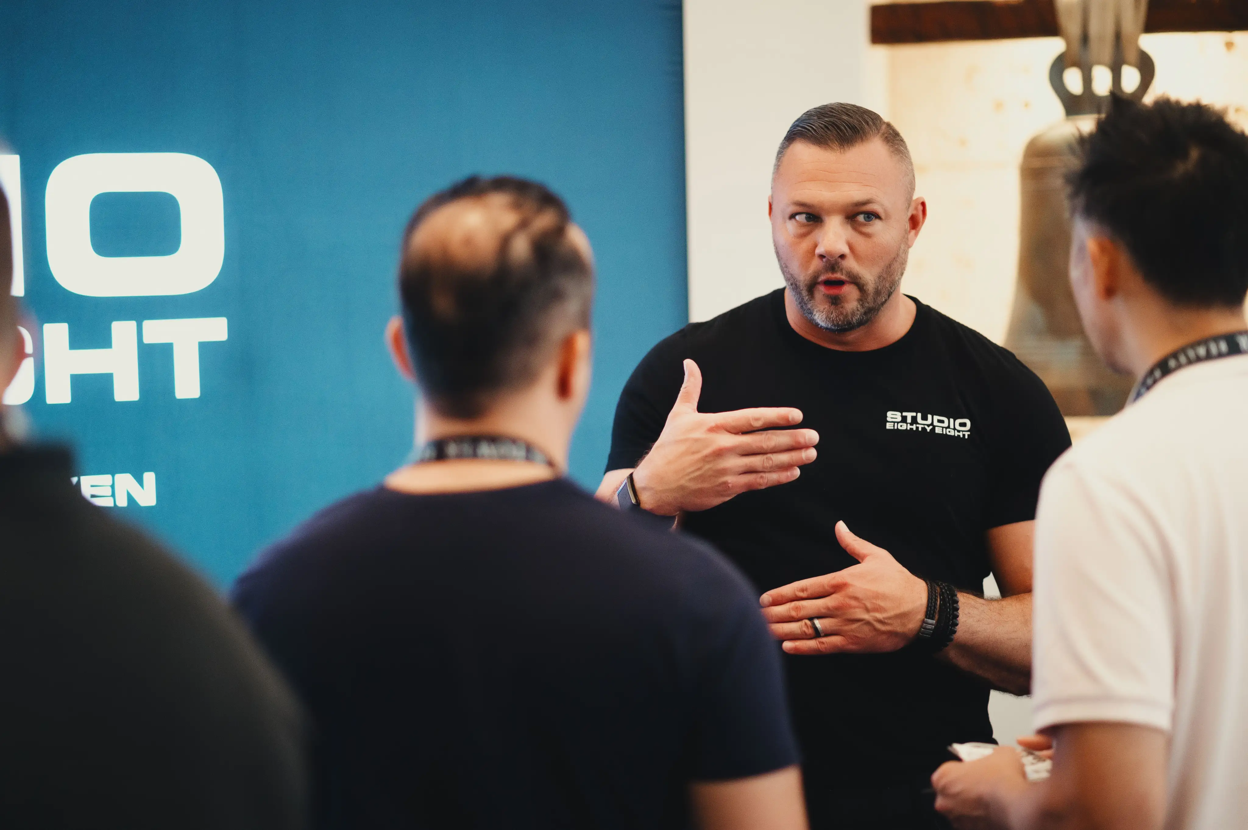 Man in black Studio Eighty Eight shirt speaks and gestures to two men listening attentively.