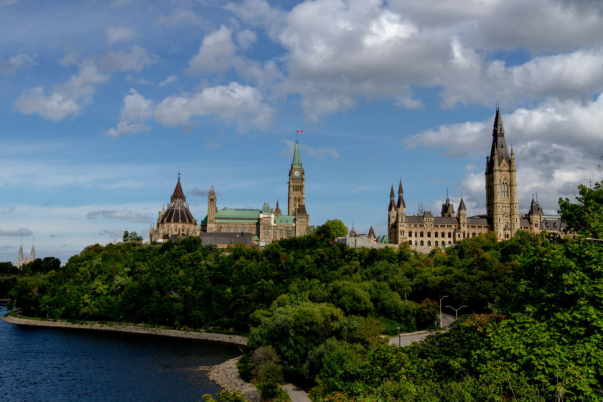 Parliament buildings atop green hill by water under a partly cloudy sky with a Canadian flag atop the central tower.