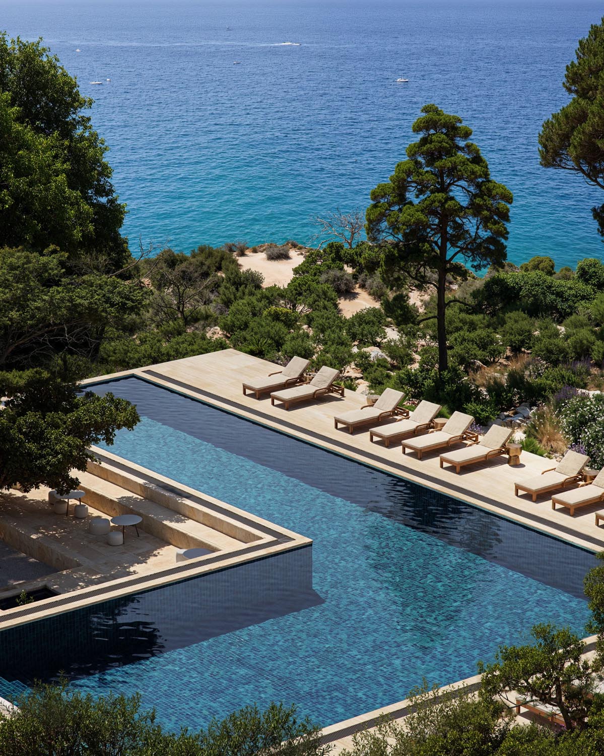 Outdoor pool with wooden loungers facing a blue sea and surrounded by lush green trees and shrubs.