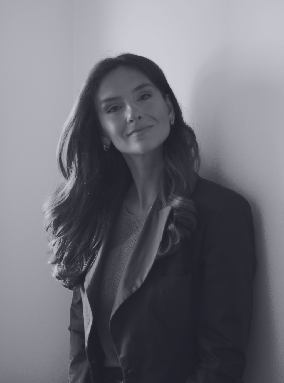 Woman with long wavy hair leaning on wall, wearing a blazer and smiling subtly.