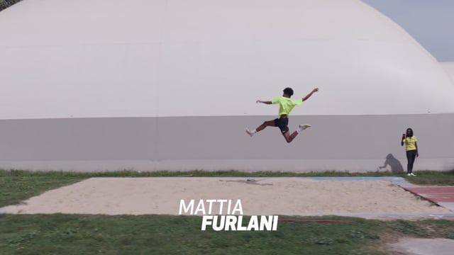 Un ragazzo in maglietta verde salta in lungo durante una gara di atletica sul campo sportivo.