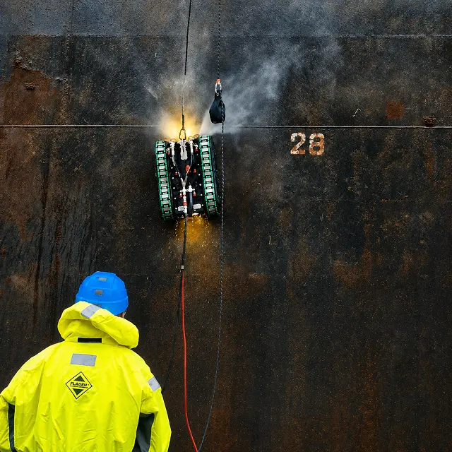 Operator in high-visibility gear controlling a hull cleaning robot from the quayside
