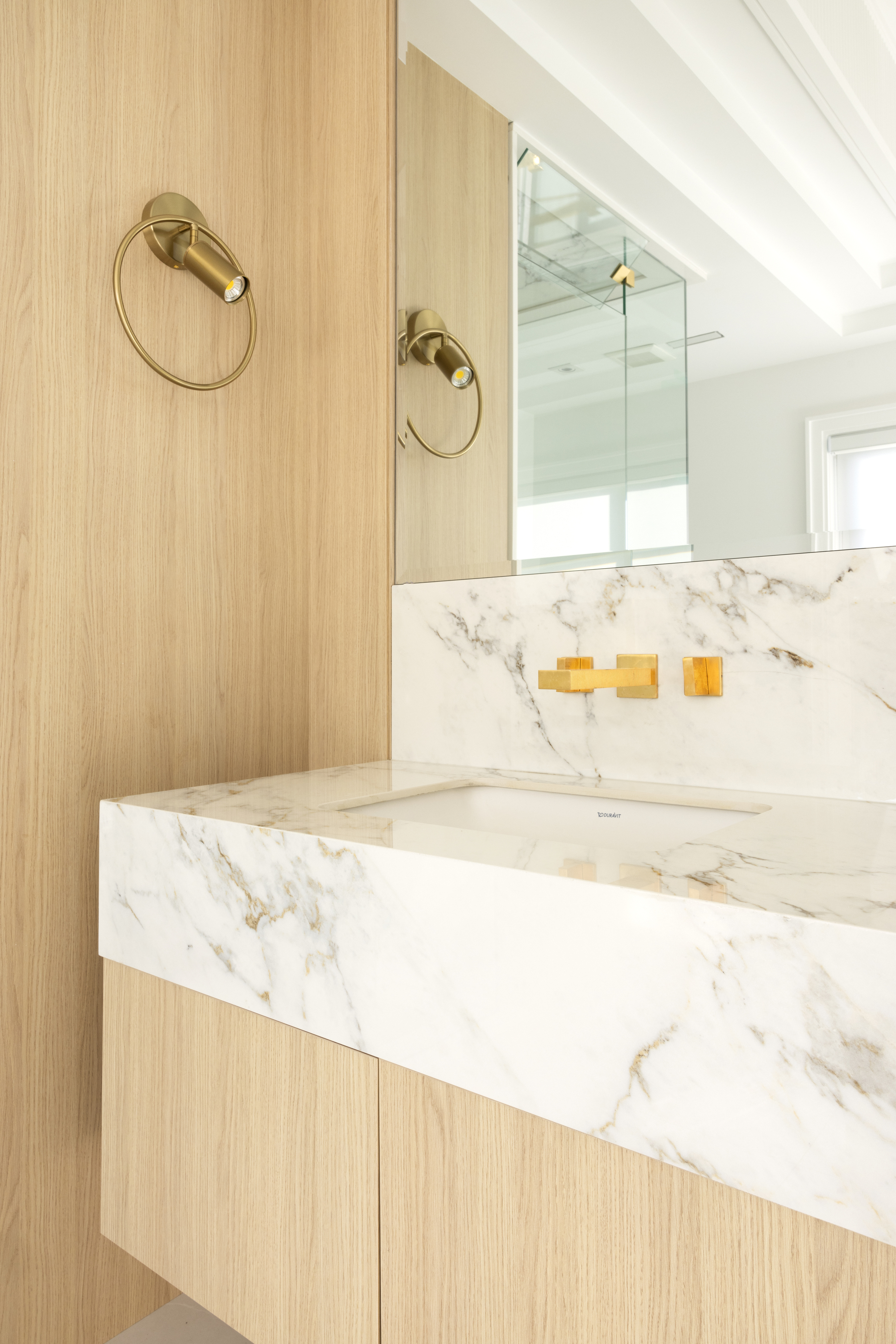 Close-up of marble bathroom sink with gold wall-mounted fixtures and wood cabinetry