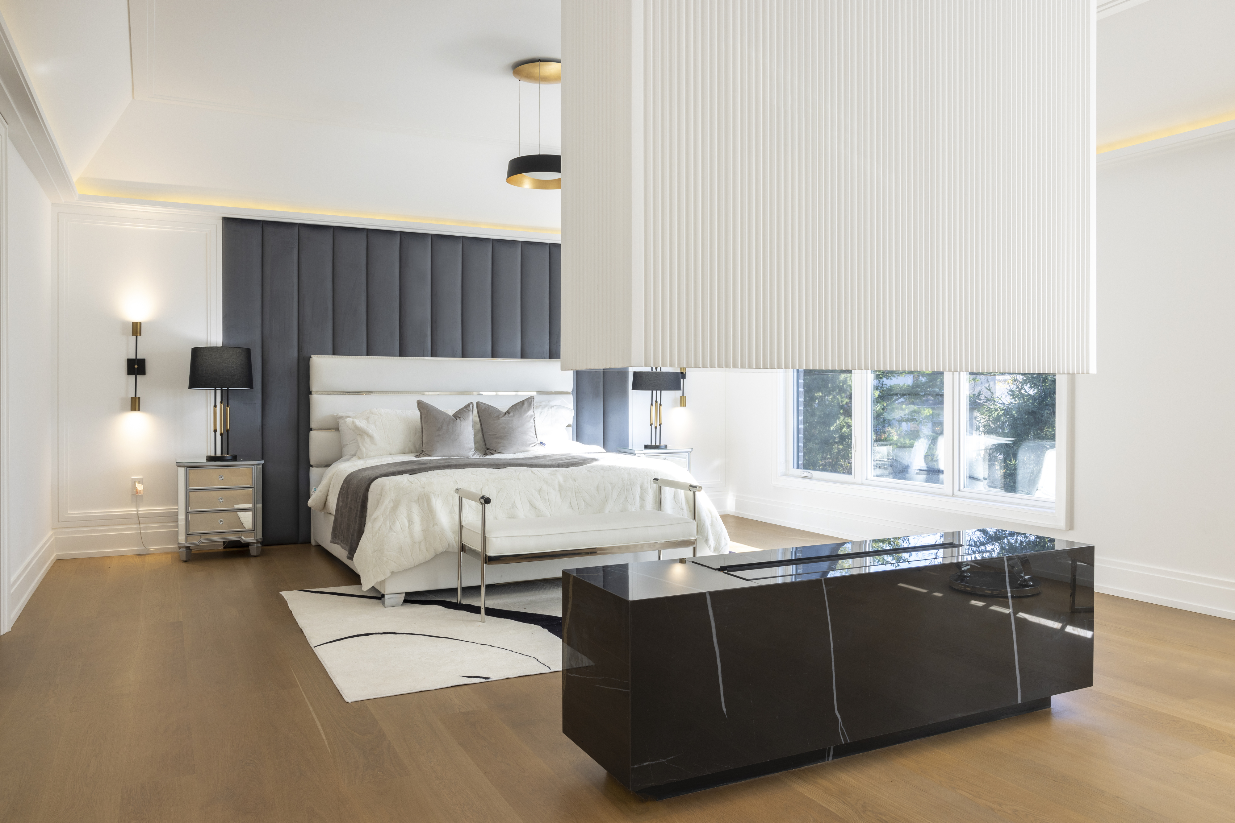 Spacious bedroom with upholstered bed, dark stone bench, and suspended light fixture