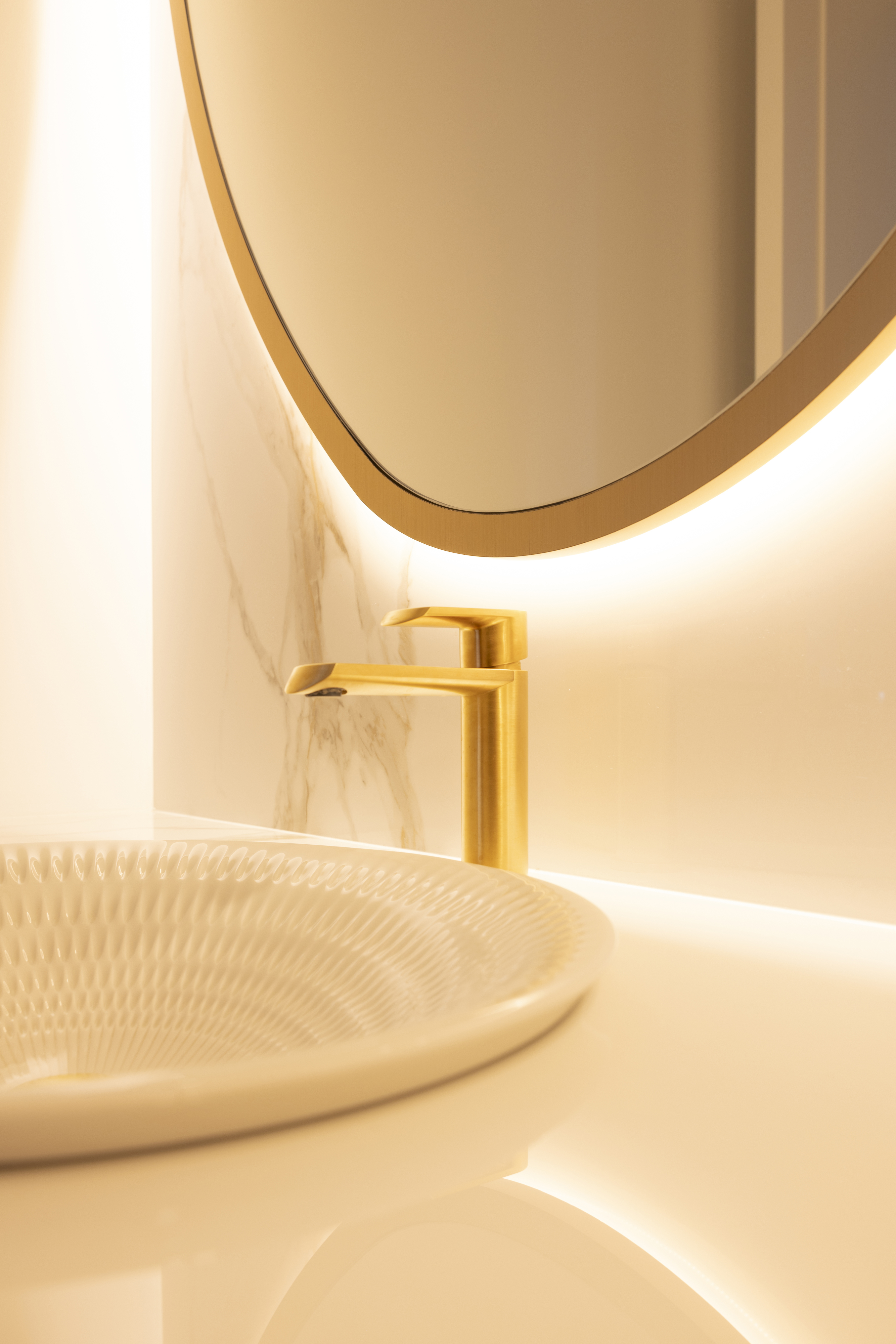 Close-up of gold faucet, textured vessel sink, and illuminated oval mirror
