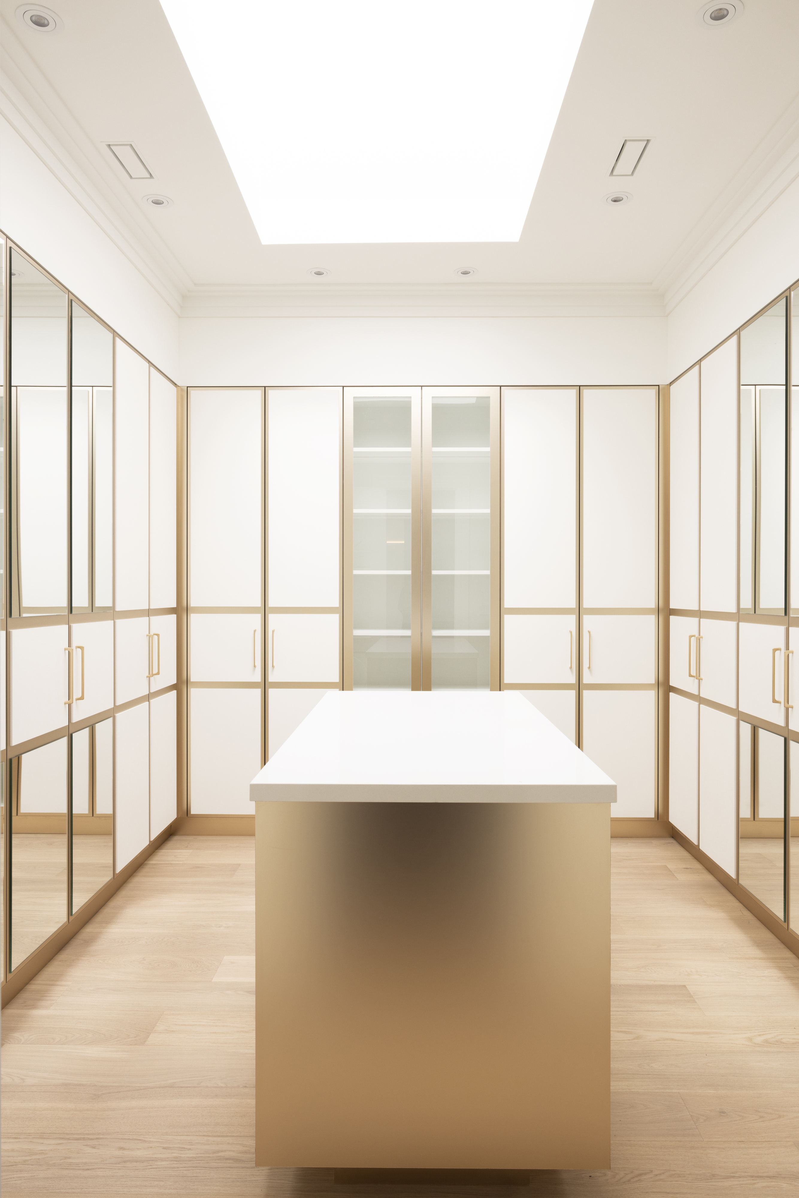 Bright walk-in closet with cream cabinetry, glass doors, gold trim, and central island