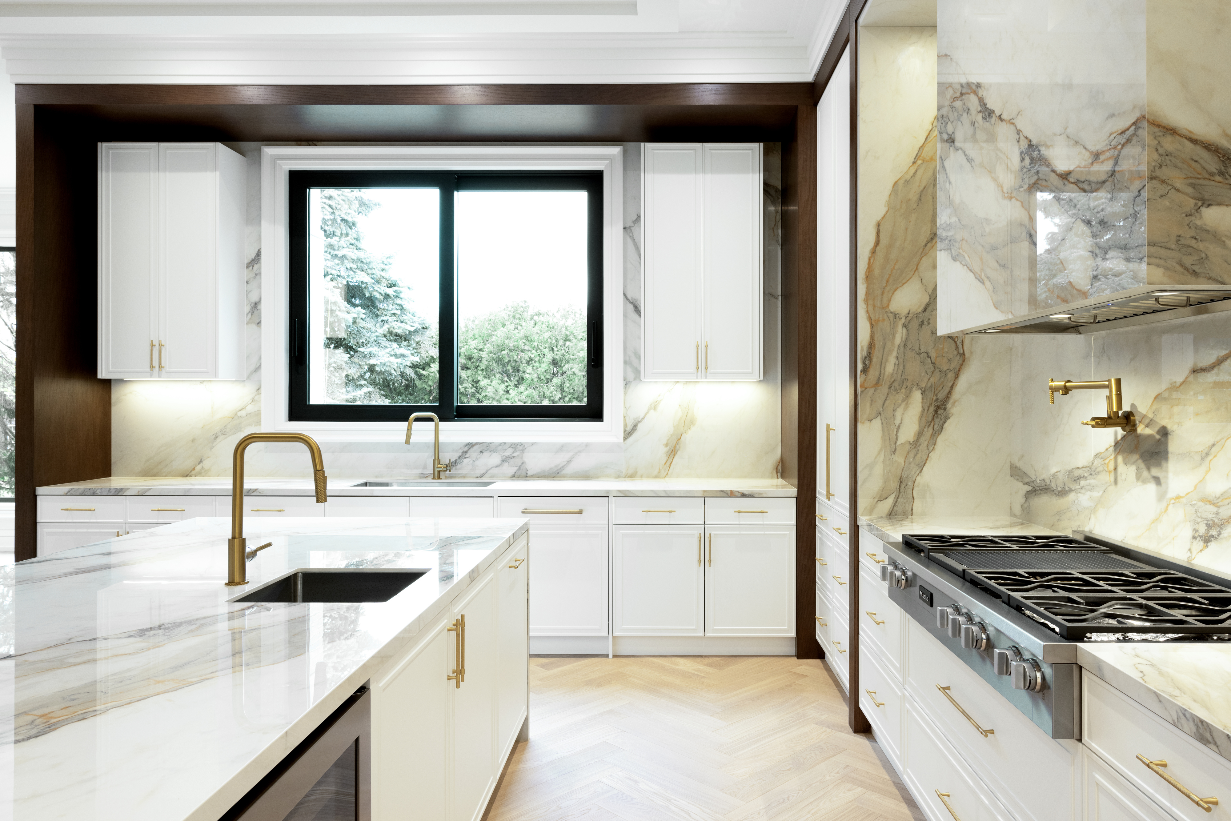 White kitchen with marble countertops, gold fixtures, dark wood trim, and large window