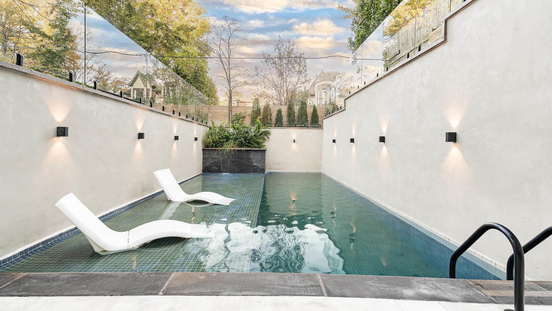 Outdoor swimming pool with submerged lounge chairs, white walls, and modern wall lighting