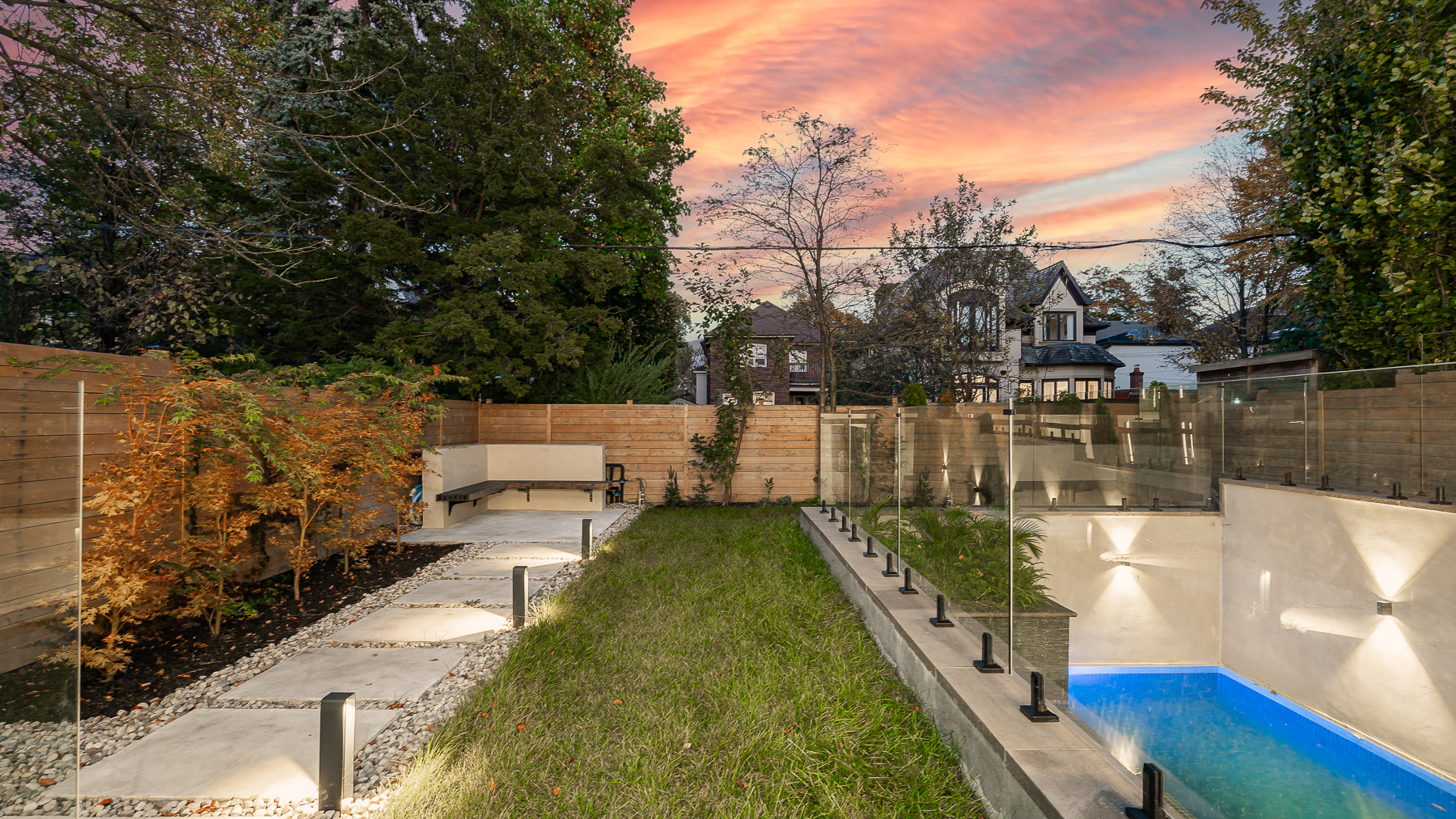 Landscaped backyard at sunset with glass railing, lawn, patio seating, and illuminated pool