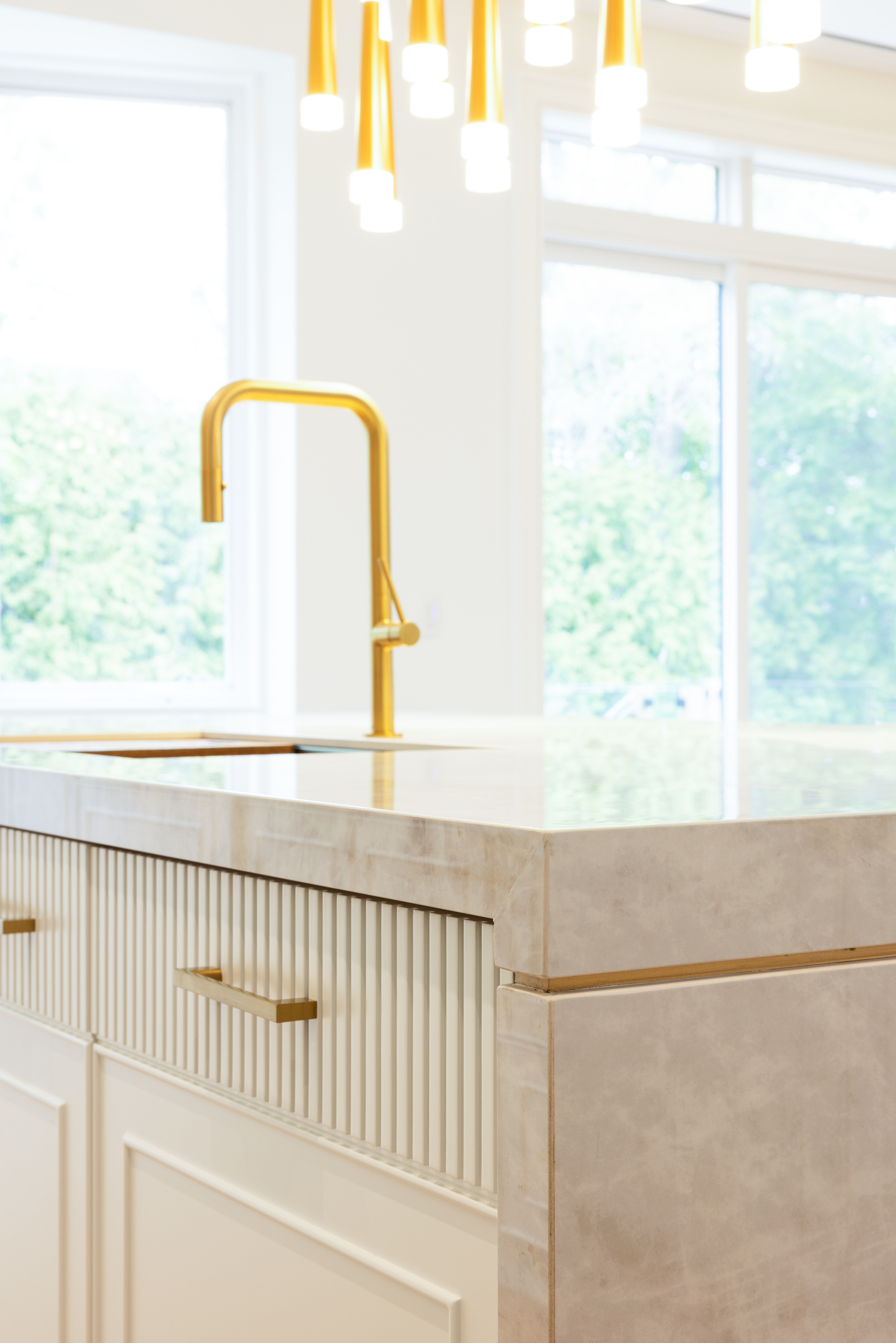 Close-up of gold kitchen faucet set into a pale stone island beside large windows