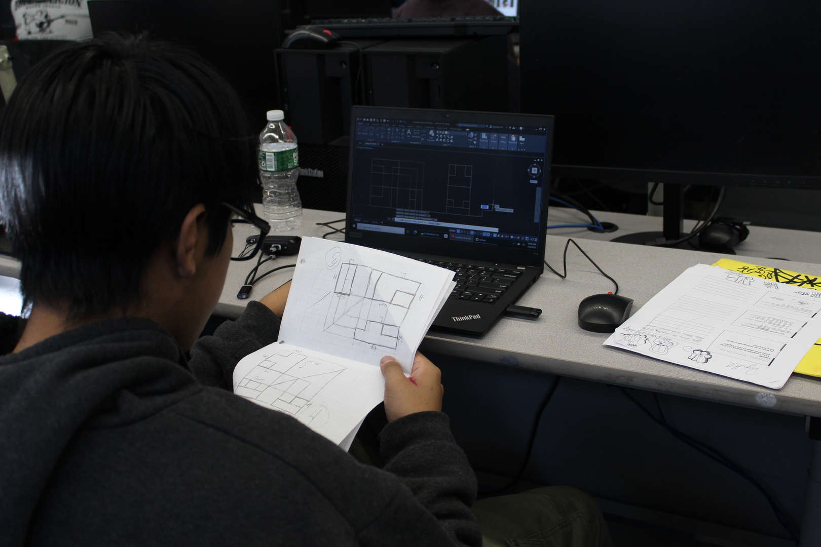 Student reviewing hand-drawn floor plan sketches beside AutoCAD laptop