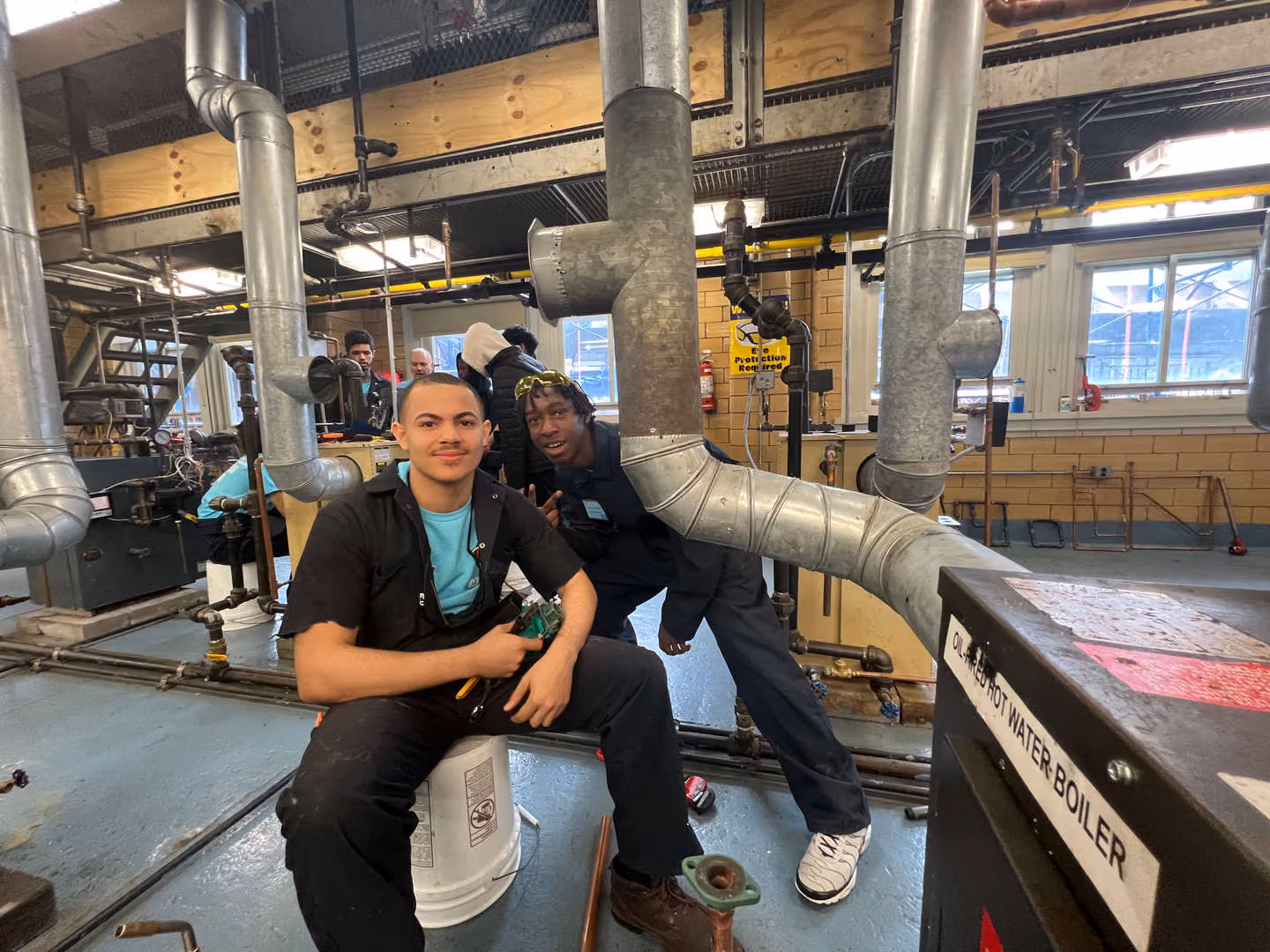 Two students posing in a vocational HVAC training workshop with ductwork