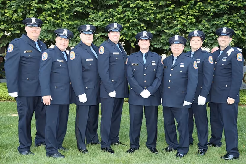 Group of nine uniformed police officers standing side by side on grass with trees behind them.