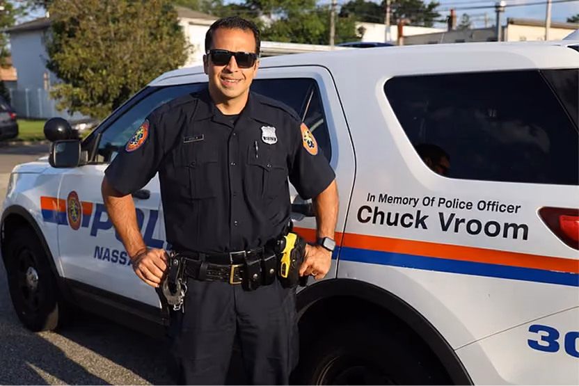 Smiling police officer in sunglasses stands in front of a police car with a memorial decal for Chuck Vroom.