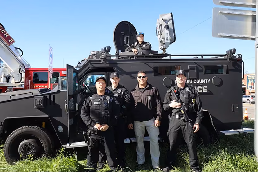 Four police officers standing in front of a black SWAT vehicle with one officer in the hatch above.