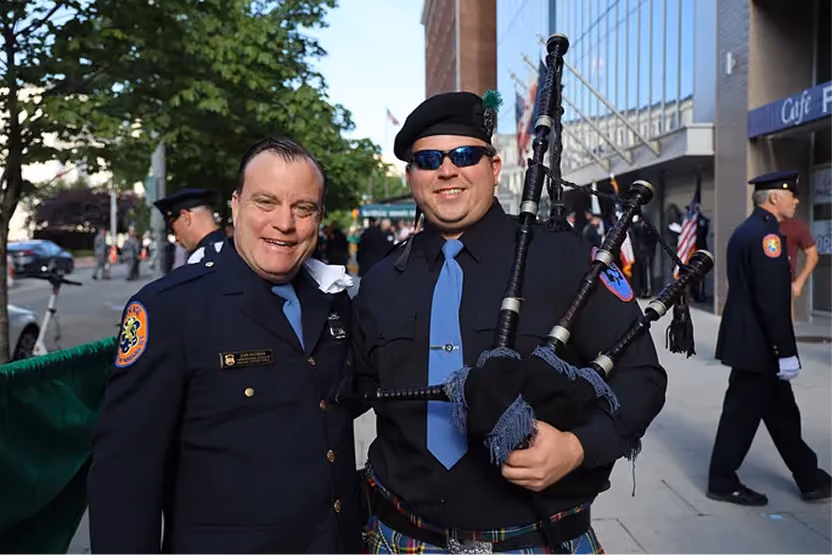 Two uniformed men smiling outdoors; one holds bagpipes, the other has a police badge.