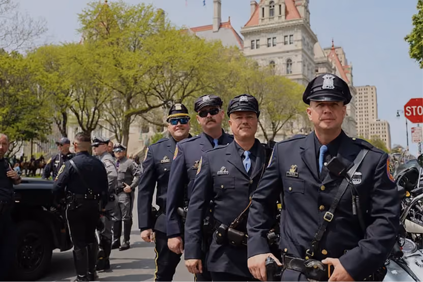 Group of police officers in uniform standing outdoors near motorcycles and buildings on a sunny day.