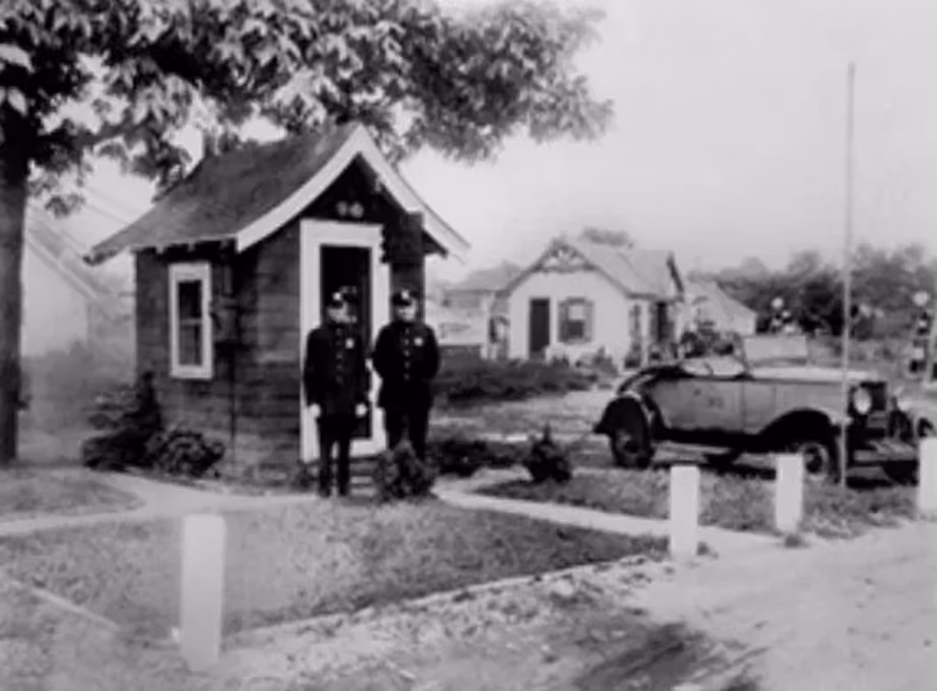 Two police officers standing outside a small guardhouse near a vintage car and houses.