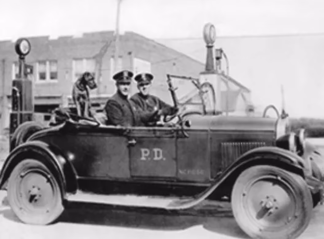 Two police officers and a dog in an old police car marked P.D. parked near gas pumps.