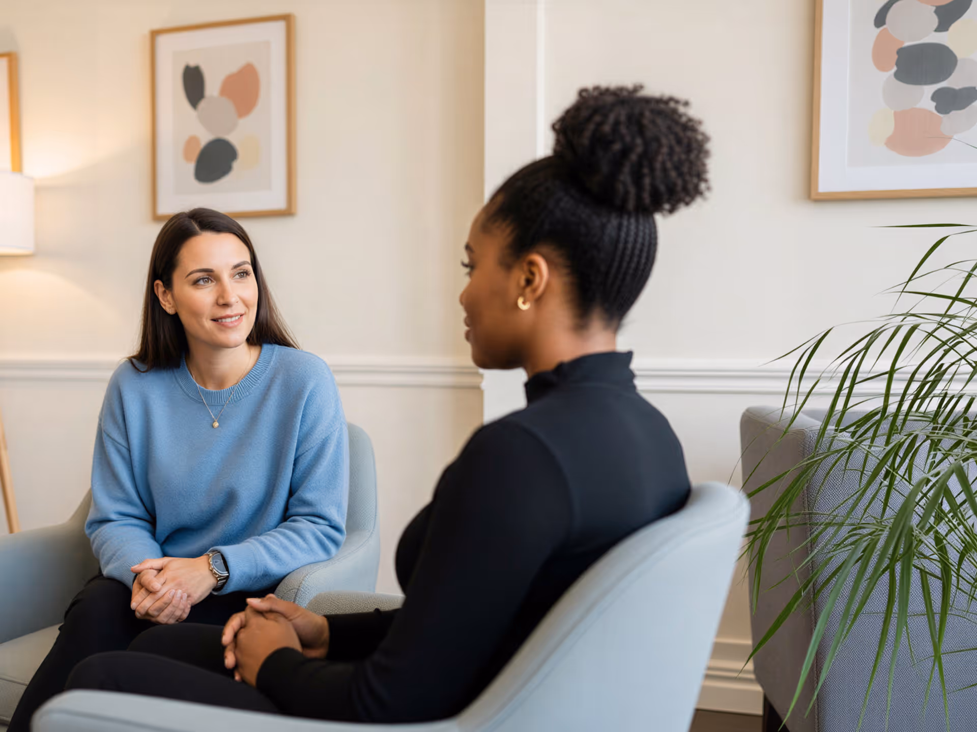 Two women sitting in armchairs facing each other and having a conversation in a cozy, softly lit room.