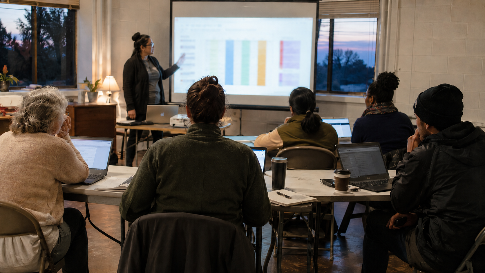A facilitator leads an evening AI training session for nonprofit staff of mixed ages working on laptops at folding tables in a modest training room.