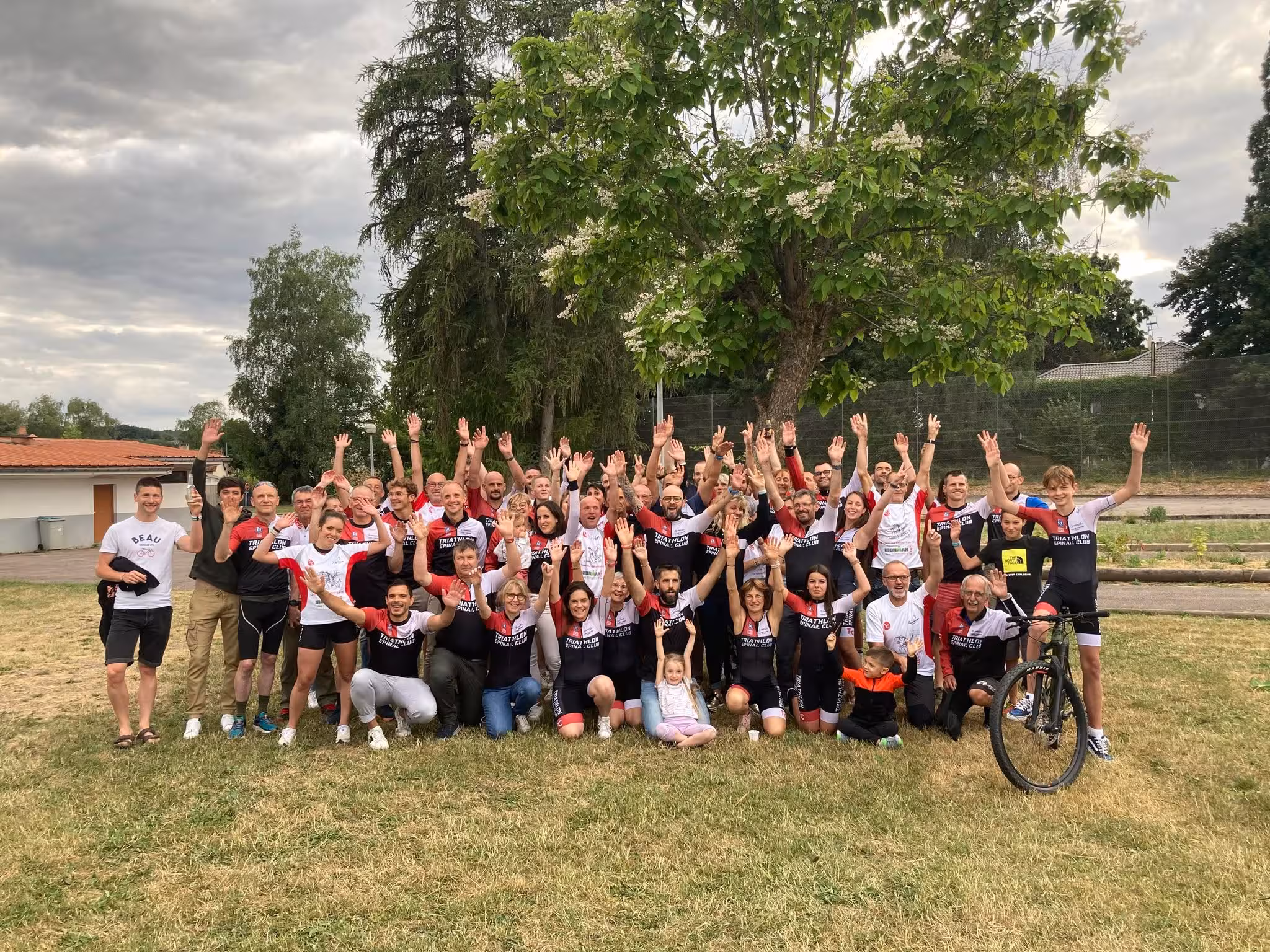 Large diverse group outdoors waving hands, some in matching sportswear with 'Triathlon Epinal Club' logo.