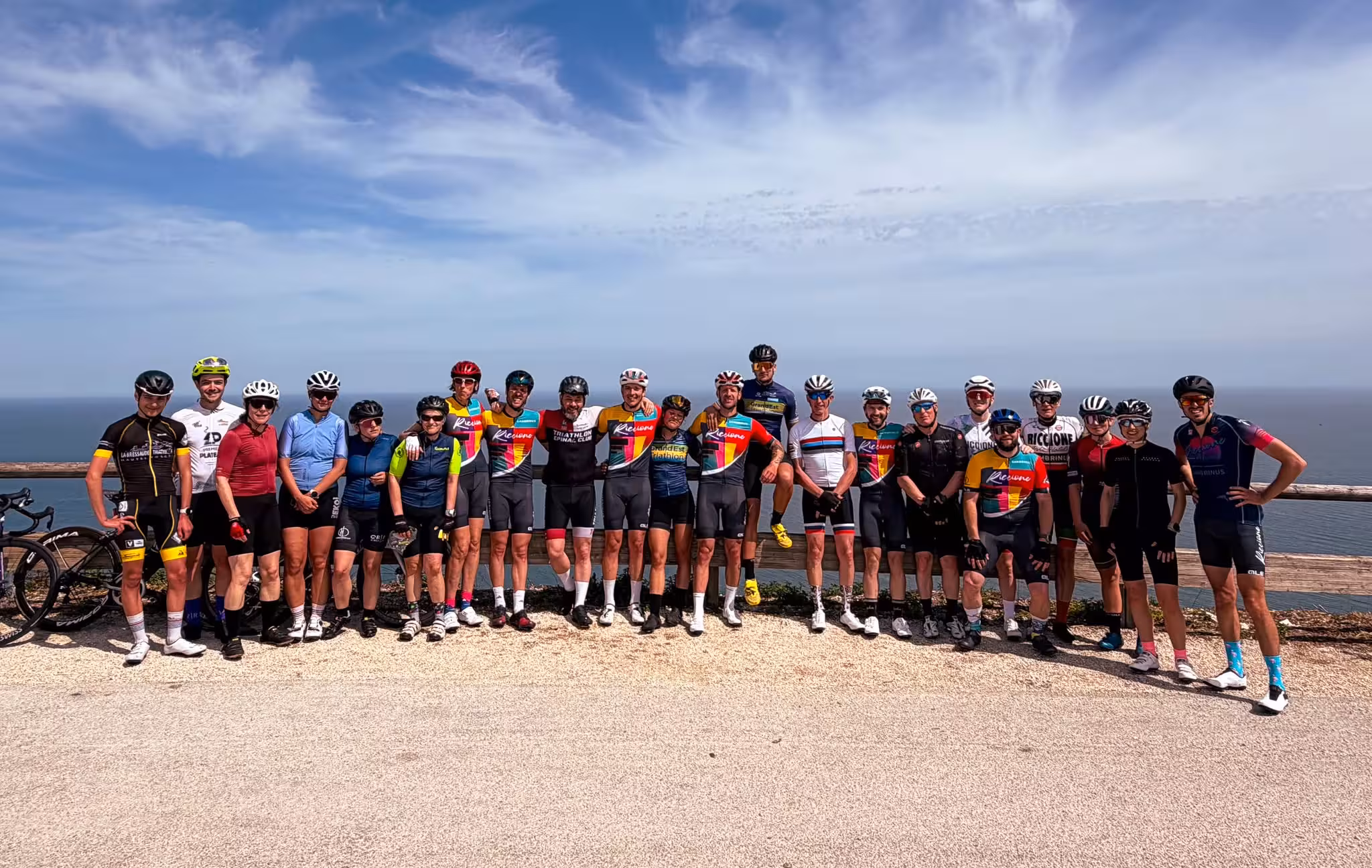 Group of cyclists in gear posing on a coastal viewpoint with blue sky and ocean behind them.