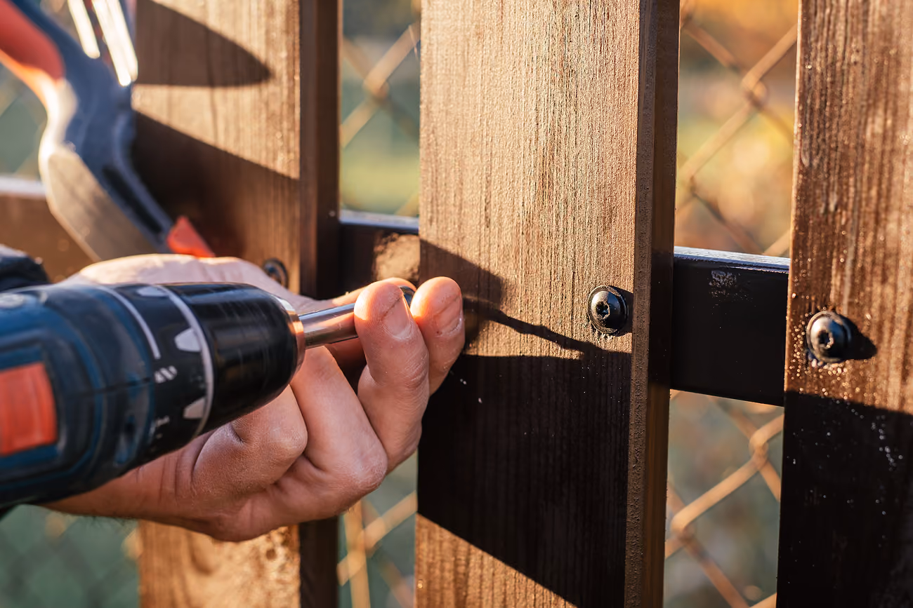 A fence builder drives a screw into a new wood fence rail in golden evening sunlight