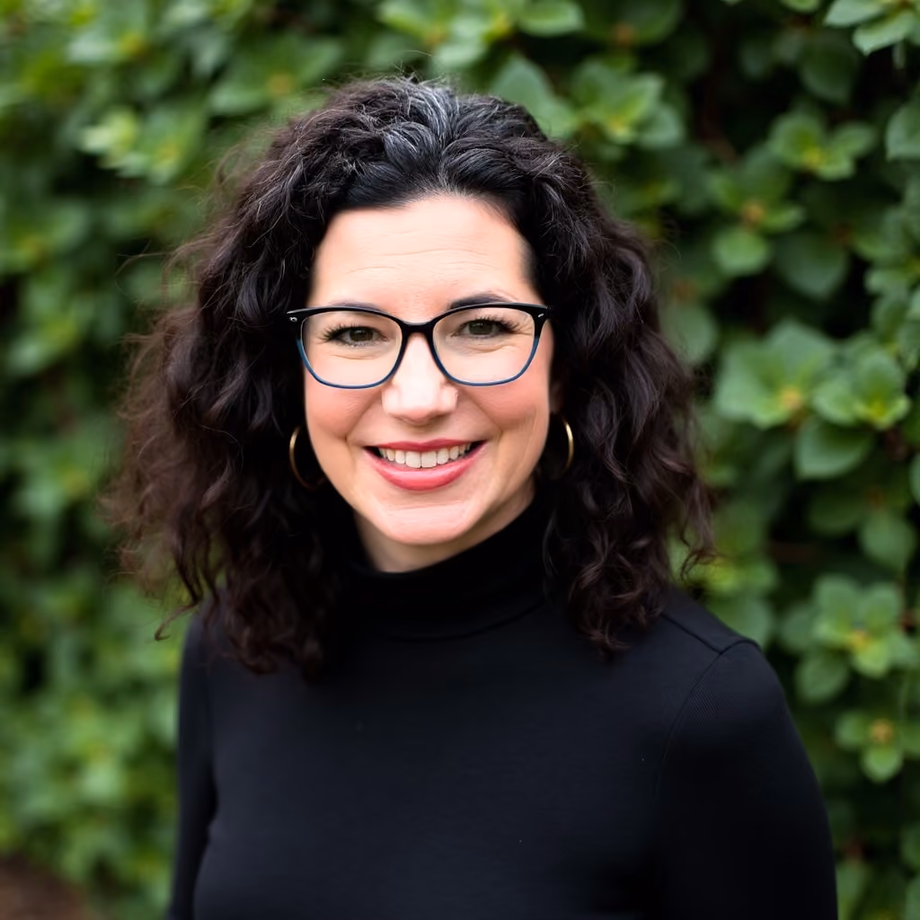 Smiling woman with curly dark hair, glasses, and hoop earrings wearing a black turtleneck outdoors.