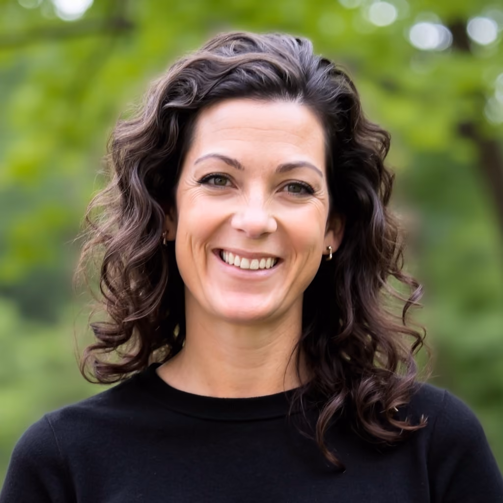 Smiling woman with curly dark hair wearing a black top, outdoors with green foliage background.