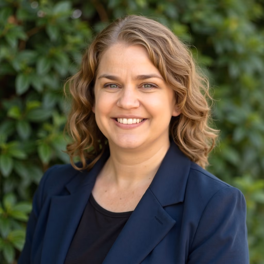 Smiling woman with curly blonde hair wearing a navy blazer against green leafy background.