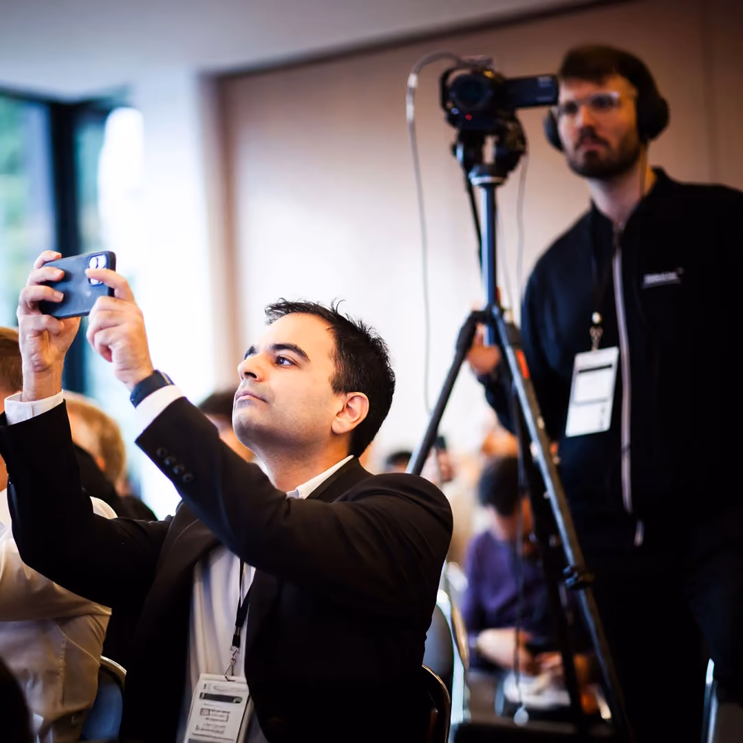 Man in suit taking a photo with phone at an event while another man records with a camera.