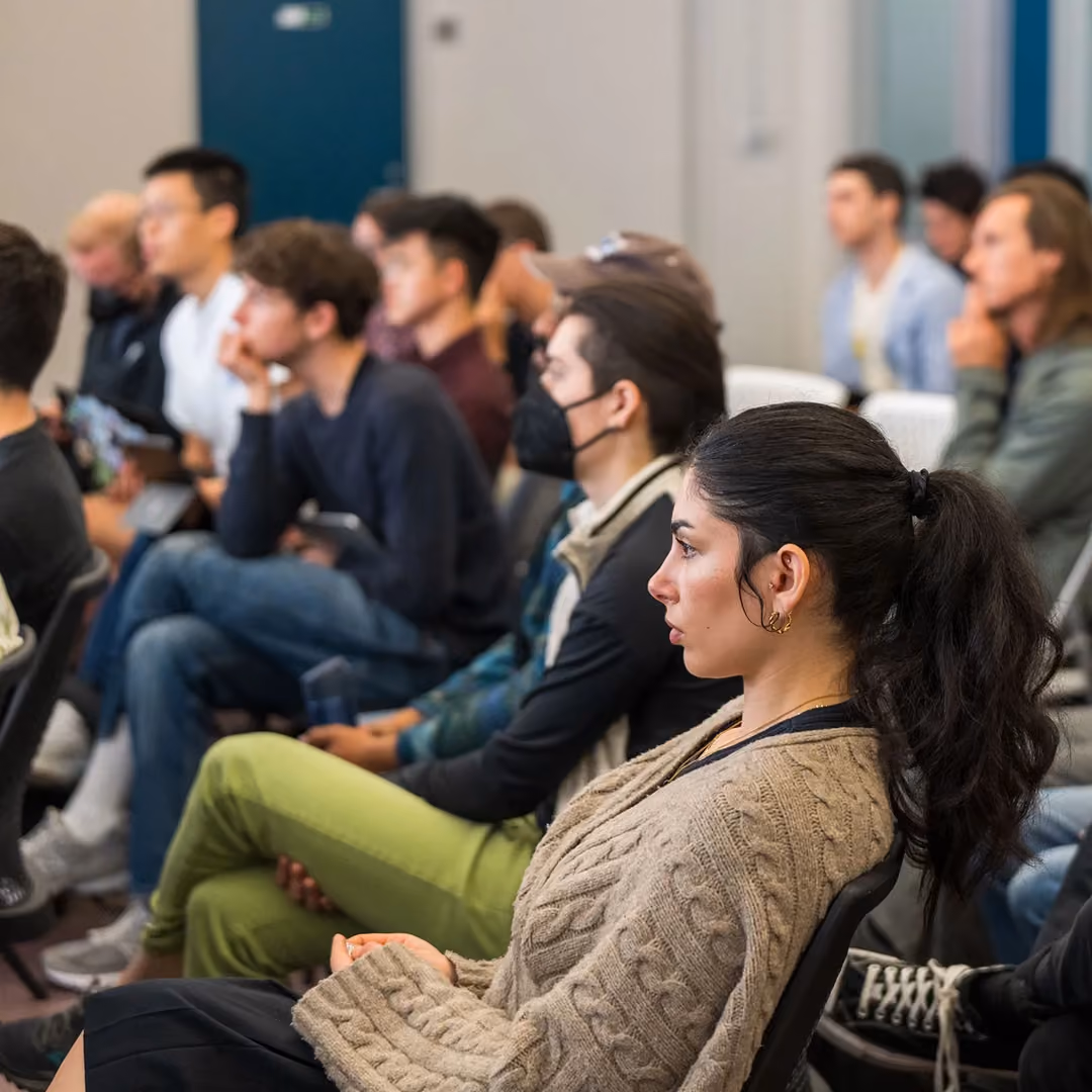 Attentive diverse group of young adults seated and listening in an indoor classroom or lecture setting.