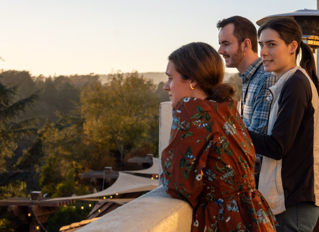 Three people leaning on a balcony railing enjoying a sunset over a forested landscape.