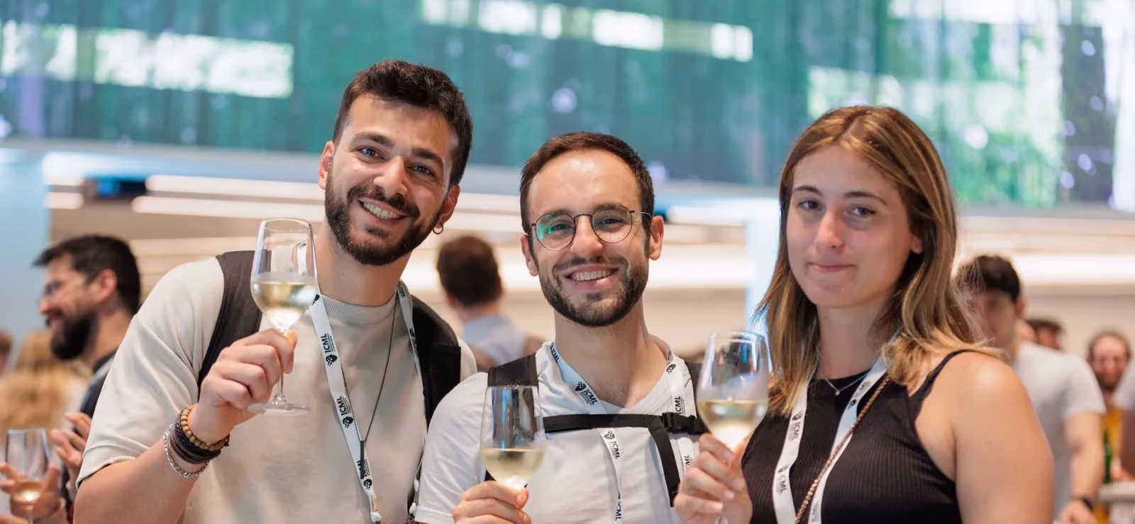 Three young adults smiling and holding wine glasses at a social event, wearing white lanyards.