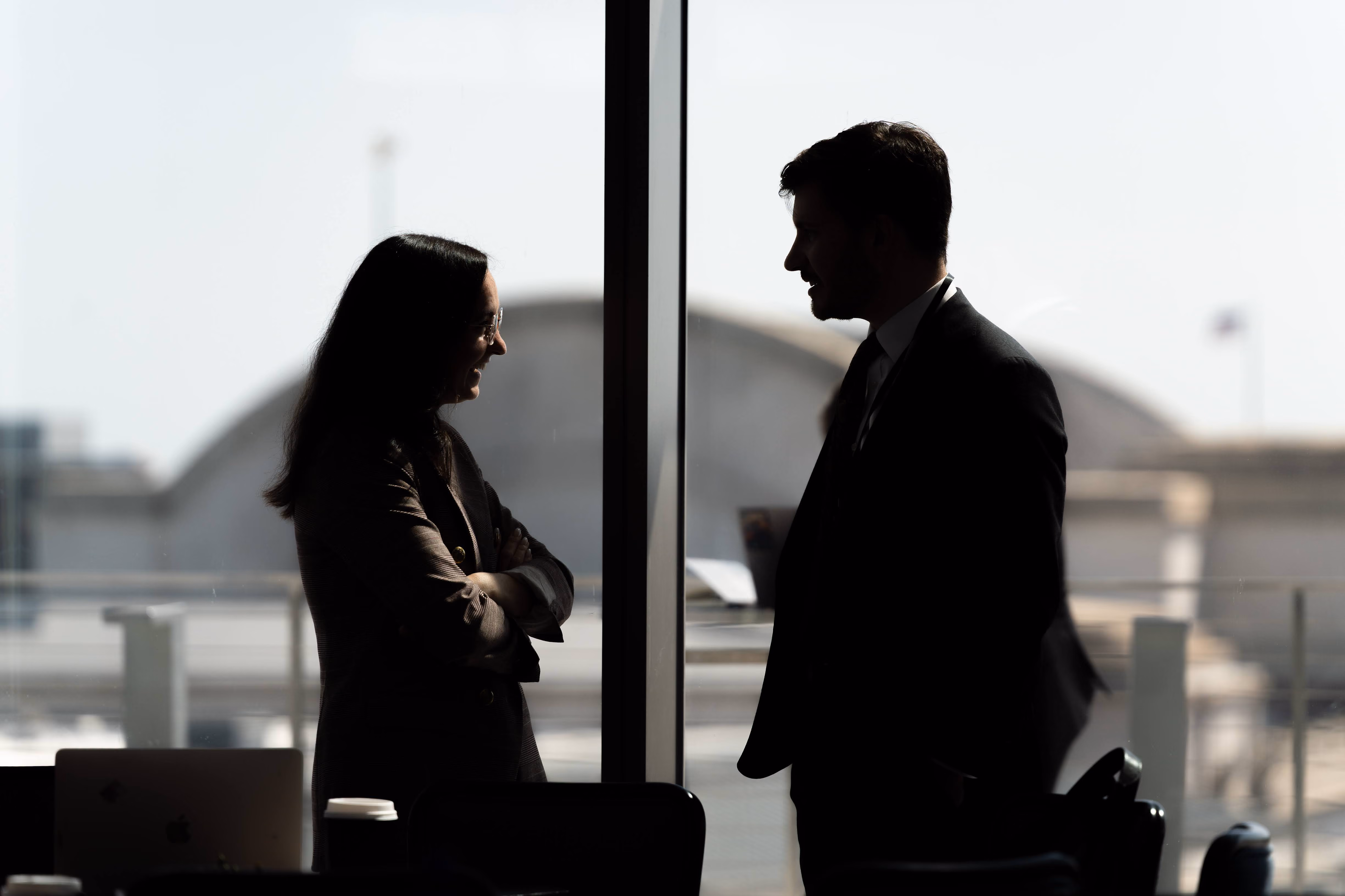 Silhouettes of two people facing each other and smiling in front of a large window with a blurred city background.
