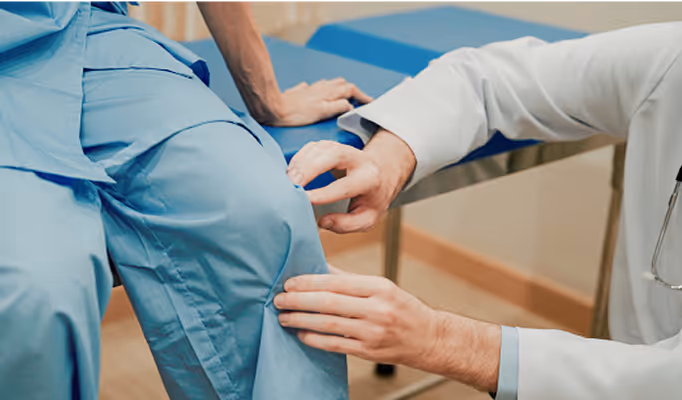 Arzt untersucht das Knie eines Patienten in blauer Kleidung in einer Klinik.