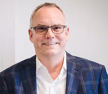 Mann mit Brille trägt ein kariertes Sakko und ein weißes Hemd vor hellem Hintergrund.