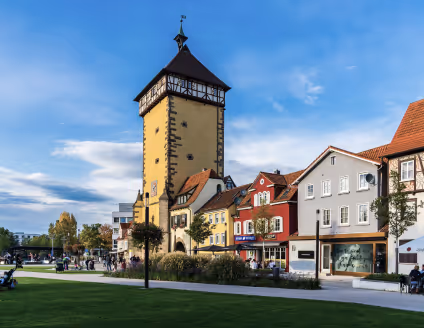 Historischer Turm neben bunten Gebäuden und einer Grünfläche bei blauem Himmel.