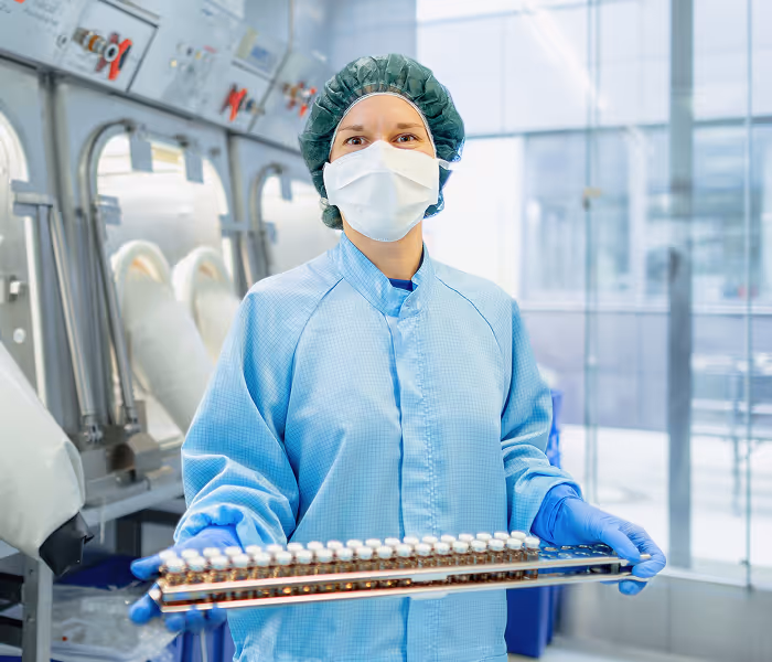 Frau in Schutzkleidung und Maske hält Tabletten auf Tablett in pharmazeutischem Reinraum.